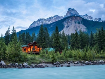 Die Lodge mit kargen Bergkipfeln im Hintergrund und türkisem Fluss im Vordergrund. Umringt von dichten Wäldern.